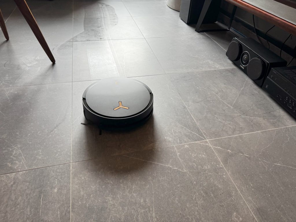 Ecovacs Deebot X8 Pro Omni robot vacuum in action, cleaning a tiled floor near a home entertainment system and wooden furniture.