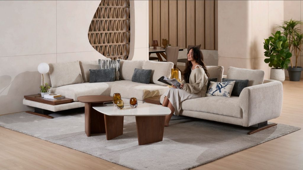 A woman in a light-colored outfit sits on a large, light grey sectional sofa in a modern living room, reading a magazine. Two nested coffee tables are in front of the sofa, which is placed on a large rug. Architectural wall features and a potted plant are visible in the background.