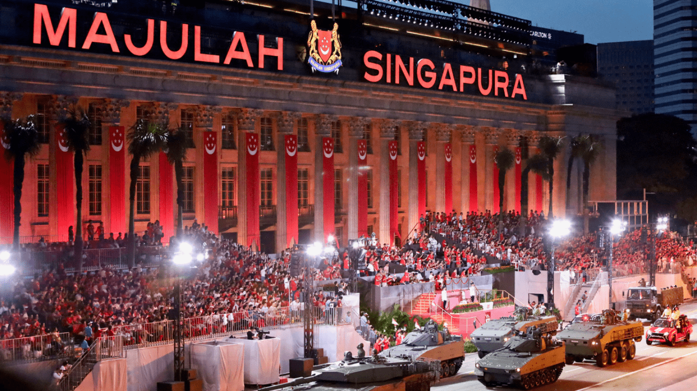 A grand night-time view of a building illuminated with red lighting, displaying the words "MAJULAH SINGAPURA" and the Singapore coat of arms. The building features large white columns adorned with vertical red and white banners. In the foreground, a large crowd of people, many wearing red and white, are gathered, looking towards the building. Below the crowd, a military parade is underway, with several armored vehicles and what appears to be a red car moving along a street. The scene is vibrant and suggests a national celebration.