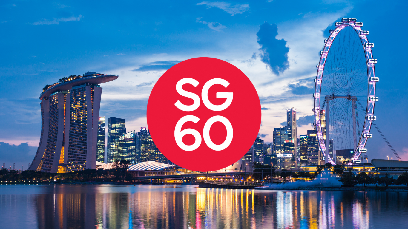 A wide shot of the Singapore skyline at dusk, featuring the Marina Bay Sands hotel on the left with its boat-like rooftop structure, and the Singapore Flyer Ferris wheel on the right, both illuminated against a deep blue sky. The city's skyscrapers reflect on the calm water in the foreground. Centered in the image is a prominent red circle with the white text "SG 60".
