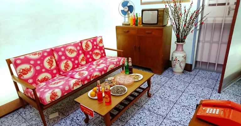A living room decorated in a retro style, possibly from the 1970s or 1980s. The room features a wooden sofa with bright pink cushions and a floral pattern. In front of the sofa is a low wooden coffee table laden with snacks and drinks, including a plate of melon seeds, nuts, and bottles of soft drinks. In the background, a wooden cabinet holds an old television set and a standing fan. A tall decorative vase with red blossoming branches sits next to the cabinet. A rotary phone is visible on a table in the foreground to the right. The floor is tiled with a grey terrazzo pattern.