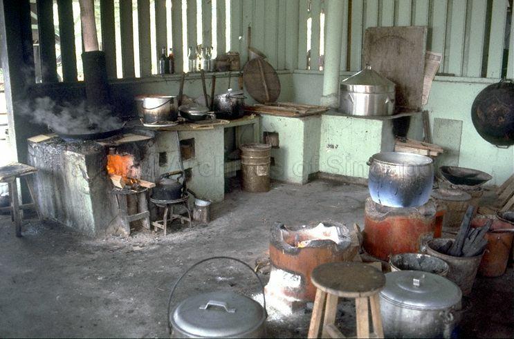 A vintage photo of a traditional Singaporean kampong kitchen. The space is rustic, with wooden walls and a concrete floor. On the left, a large, rectangular wood-fired stove is in use, with smoke rising from a large wok. A smaller stove with a kettle sits next to it. Various pots, pans, and utensils are scattered around the kitchen, including a large steamer pot, a hanging wok, and multiple aluminum pots on the floor. Several round wooden stools are placed throughout the scene.