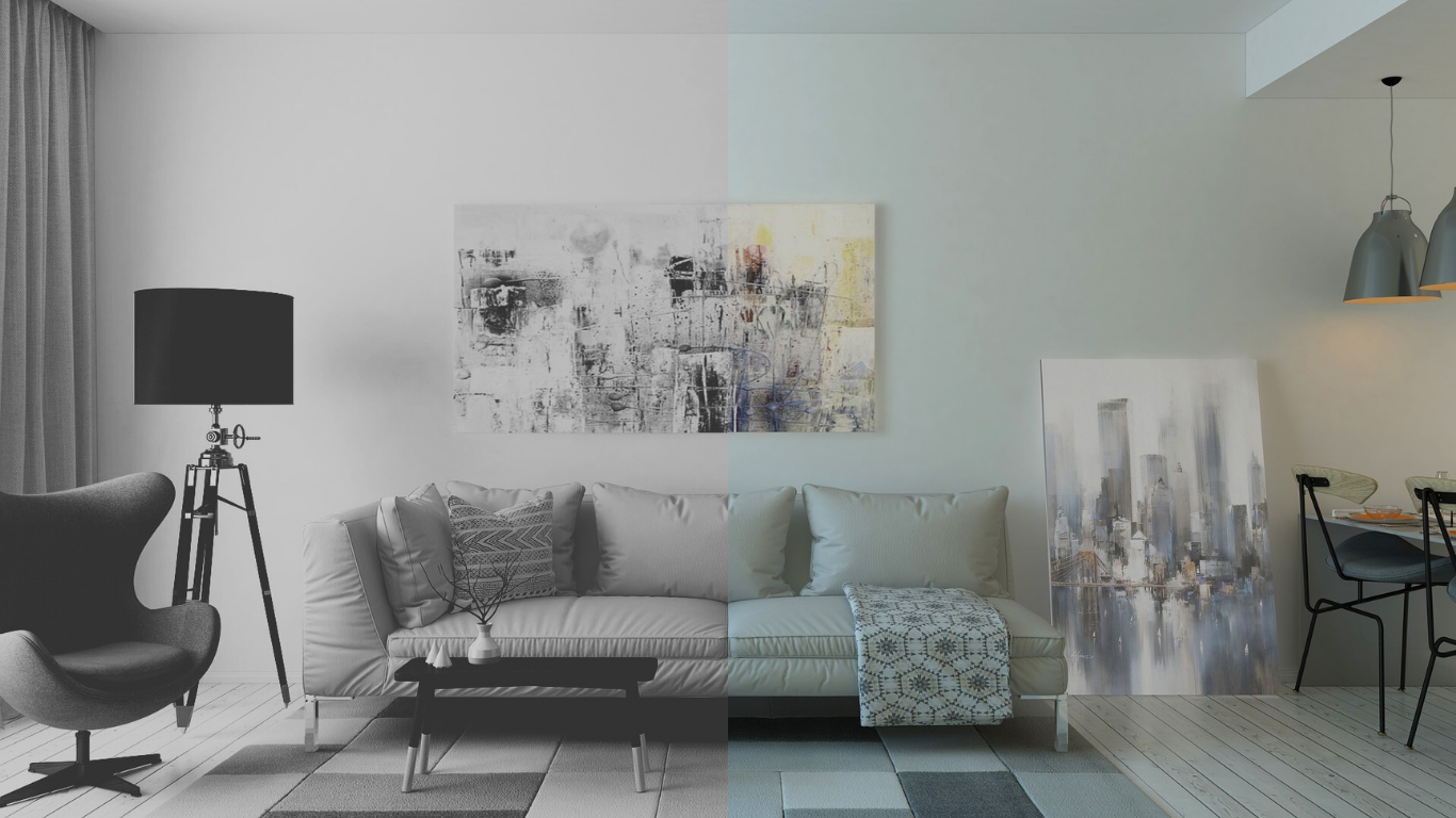 A stylish and modern living room and dining area, primarily in grayscale, with a few subtle touches of colour. The living room features a light grey sofa with several cushions, a throw blanket with a geometric pattern, a black coffee table, and a large abstract painting with hints of yellow and blue on the wall behind it. To the left is a dark grey wingback chair and a black tripod floor lamp with a drum shade. The dining area on the right has a black table, chairs, and two hanging pendant lamps. A second painting of a city skyline rests against the wall in the dining area. The floor is covered with a striped rug and light wooden planks.