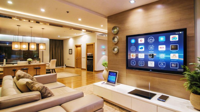 A spacious, modern open-plan living and dining area with a focus on smart home technology. The living room features a large sectional sofa and a white, low-profile media console beneath a wall-mounted smart TV that displays an app interface. A tablet is propped up on the console, and a smartphone and other devices are also visible. The TV wall is covered in a light wood-grain panel, with three circular decorative panels to its left. The adjacent dining area features a kitchen island with bar stools and hanging pendant lights with wicker shades. The room has light wood flooring and a mix of recessed and accent lighting.