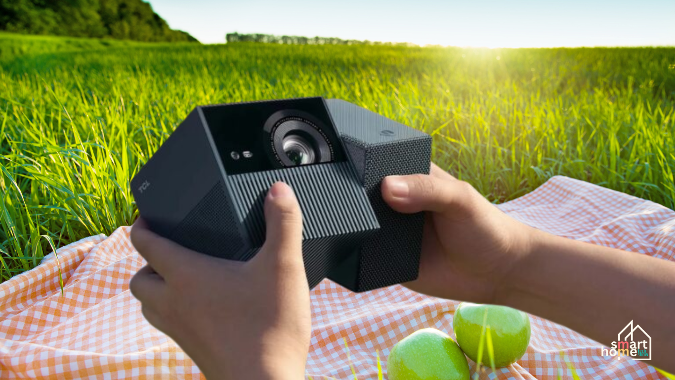 A pair of hands holds a black TCL PlayCube projector over a red and white checkered picnic blanket. The projector has a unique hexagonal shape with a prominent lens on its front face. Two green apples rest on the blanket next to the projector. In the background, a person is seen enjoying a picnic in a sun-drenched, grassy field under a clear blue sky. The image has a watermark in the bottom right corner that says "Smart Home.
