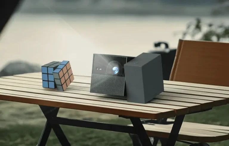 A TCL PlayCube projector sits on a wooden slatted picnic table. The projector has a two-part design, with a main cubic unit containing the lens and a second detachable cube that serves as a speaker and power bank. A Rubik's Cube with blue, red, and orange faces is next to the projector. In the blurry background, there is a body of water and a camping chair.