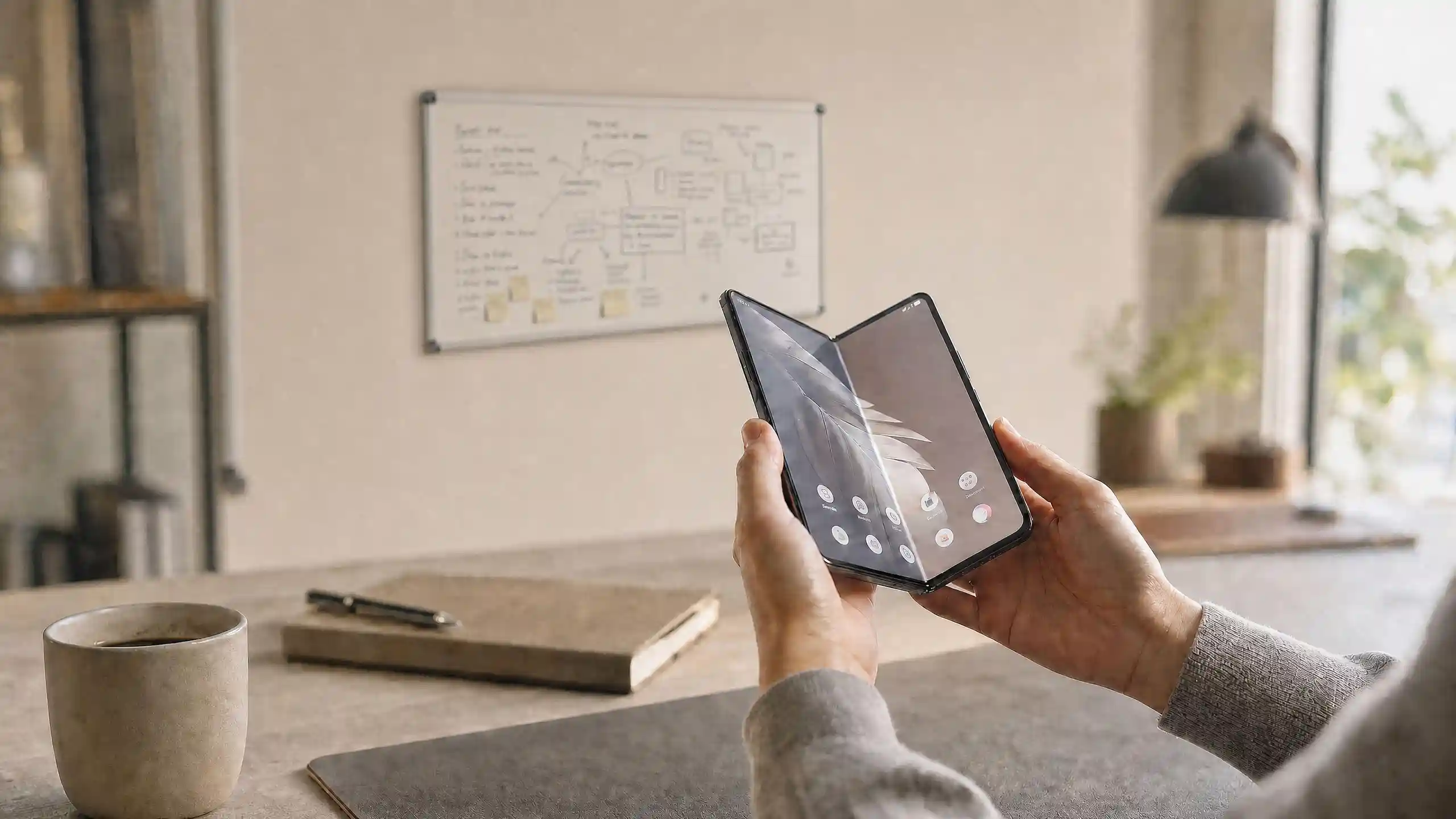 Hands unfolding a book-style foldable smartphone in a bright, minimalist office, with a whiteboard and coffee mug visible in the background.